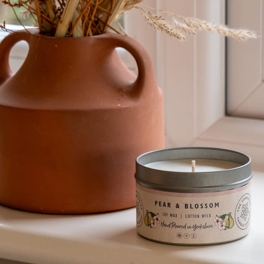 Candle labeled 'Pear & Blossom' next to a terracotta vase with dried plants on a windowsill.