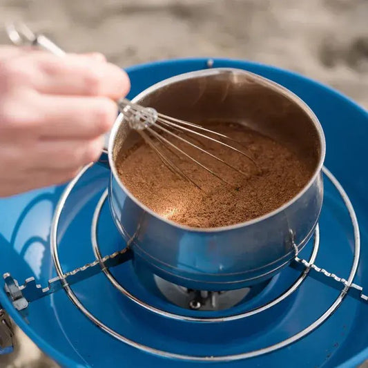 Person stirring a pot of hot chocolate on a camping stove with a whisk.