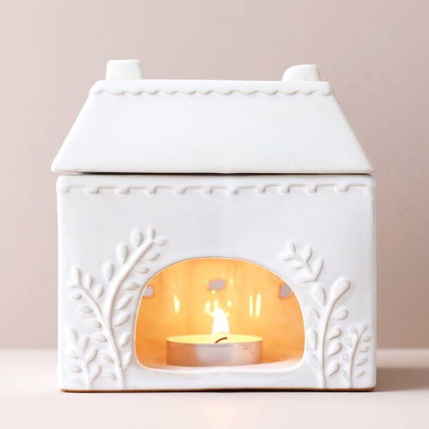 White ceramic house-shaped lantern with a lit candle inside on a light pink background