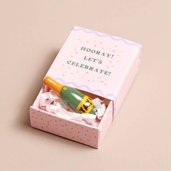 Small bottle with a colorful cap in a pink box with 'Hooray! Let's Celebrate!' text on a beige background