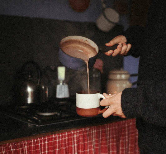 Person pouring hot chocolate into a mug with steam in a cozy kitchen setting.