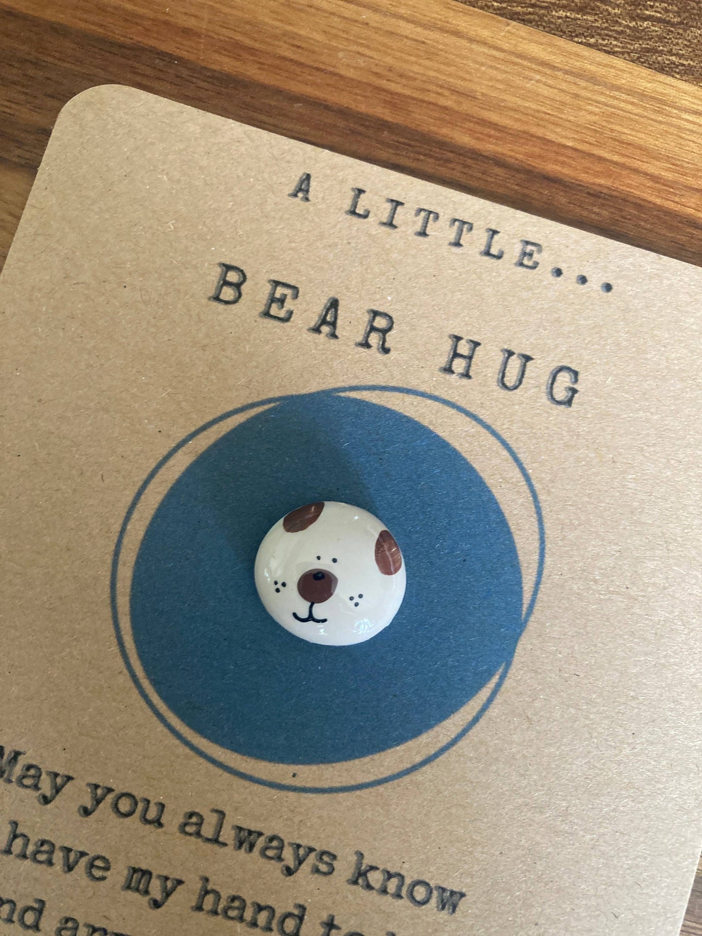 Card with a pebble featuring a bear face on a blue background, placed on a wooden surface.