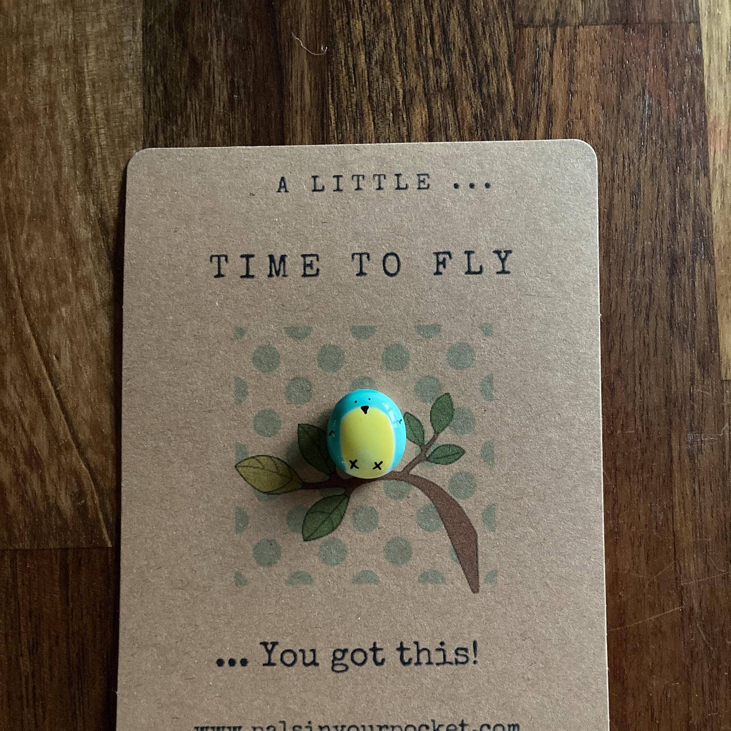 Small figurine of a bird on a branch with a motivational message on a brown card, placed on a wooden surface.