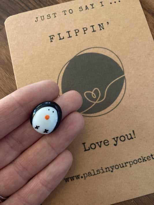 Small round black and white penguin charm held in a hand with a branded card in the background.