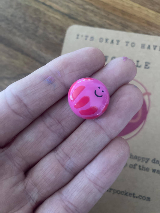 Pink charm with smiley face held between fingers on a wooden surface