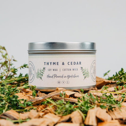 Candle in a tin labeled 'Thyme & Cedar' surrounded by herbs on a light background