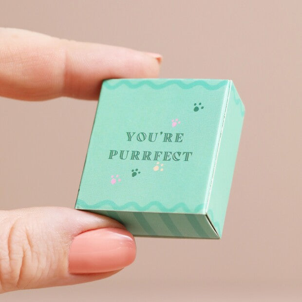 Small green box with 'YOU'RE PURRFECT' text held between fingers against a beige background