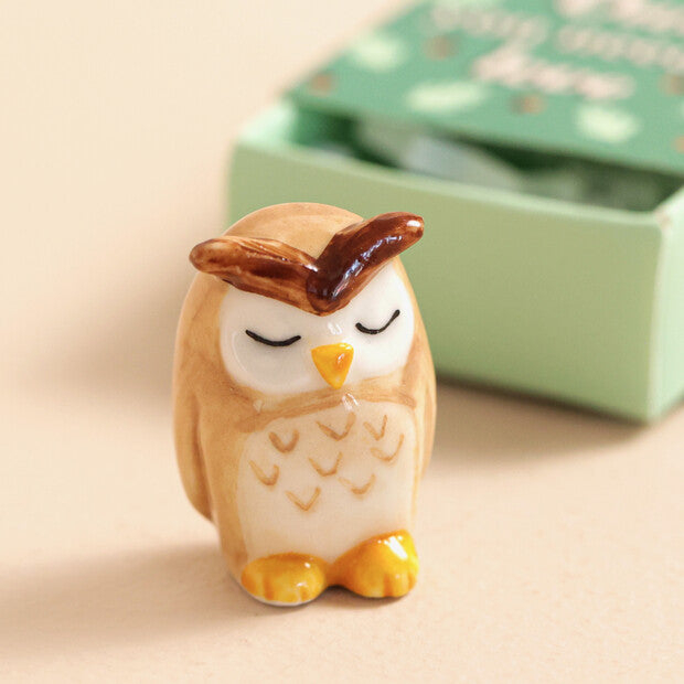Small owl figurine with closed eyes on a beige surface, with a green box in the background.