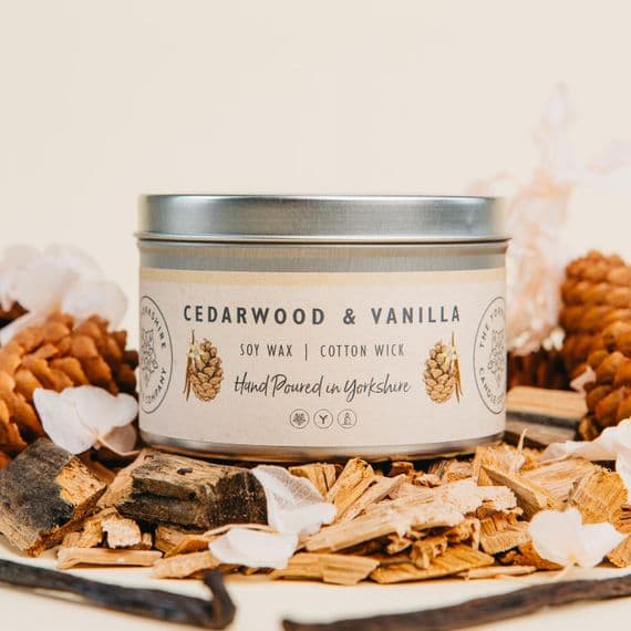 Candle tin labeled 'Cedarwood & Vanilla' surrounded by wood chips and pine cones on a light background