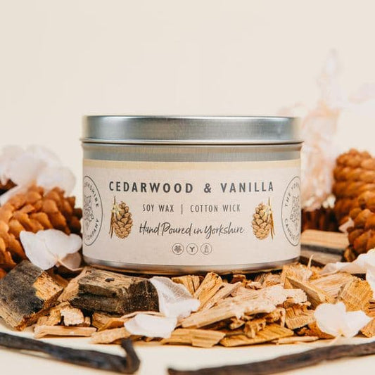 Candle tin labeled 'Cedarwood & Vanilla' surrounded by wood chips and pine cones on a light background