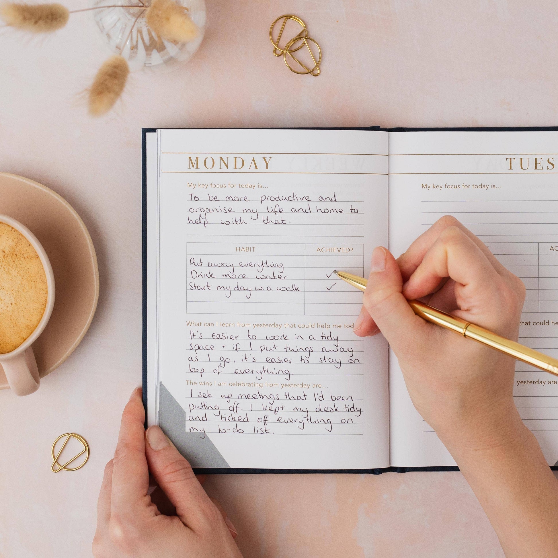 Person writing in a planner with a cup of coffee and decorative elements on a light pink surface