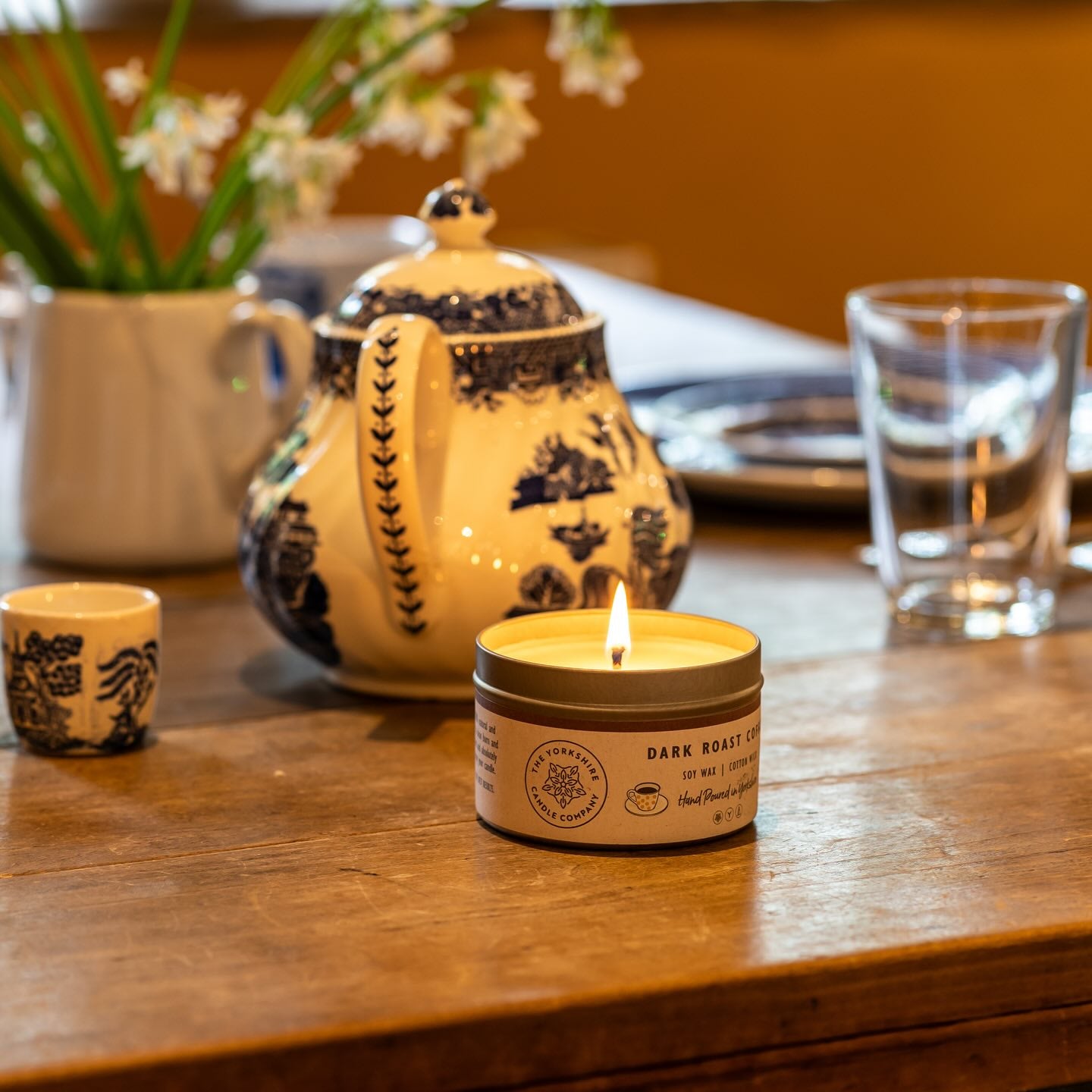Candle with a decorative label on a wooden table with a teapot and flowers in the background.