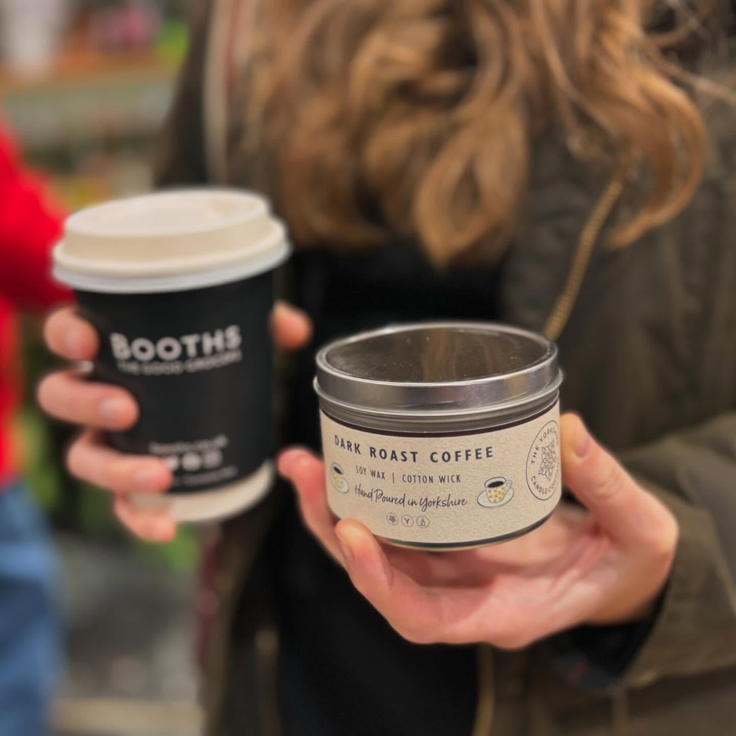 Person holding a coffee cup and a candle labeled 'Booths' and 'Dark Roast Coffee'.