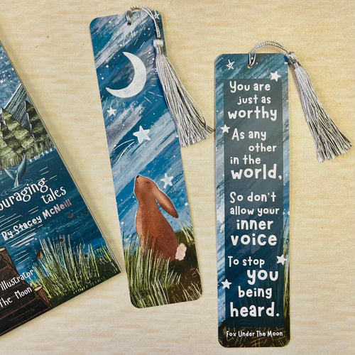 Two bookmarks with tassels on a wooden surface, one featuring a moon and stars design, the other with a motivational quote.