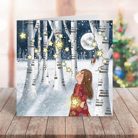Card with a winter scene of a girl in a snowy forest with stars and a moon, on a wooden surface.