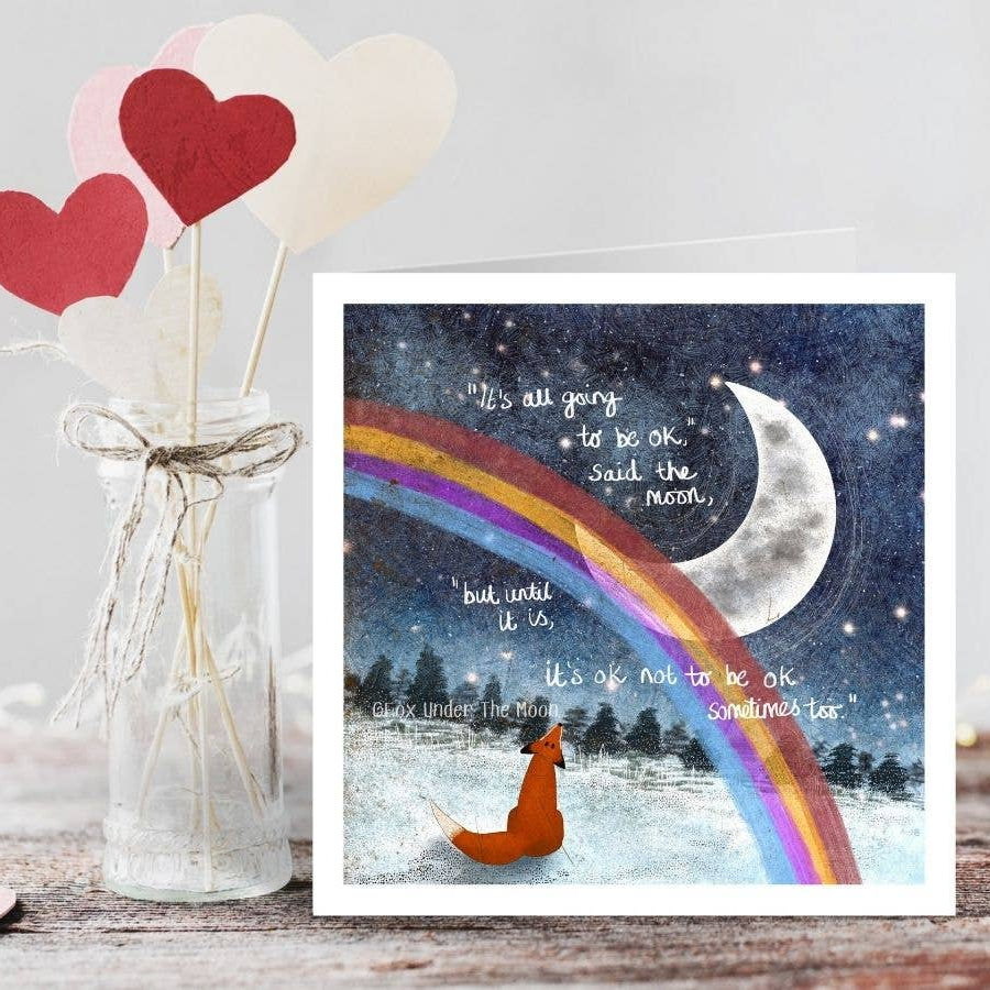 Decorative card with a fox and rainbow next to a vase with heart-shaped objects on a wooden surface.