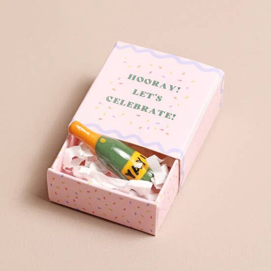 Small bottle with a colorful cap in a pink box with 'Hooray! Let's Celebrate!' text on a beige background