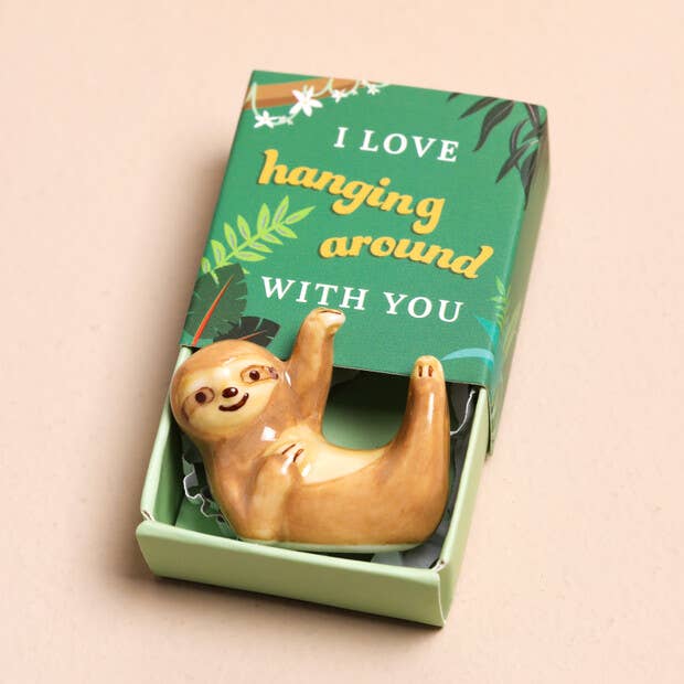 Sloth figurine in a box with a green lid featuring text on a beige background