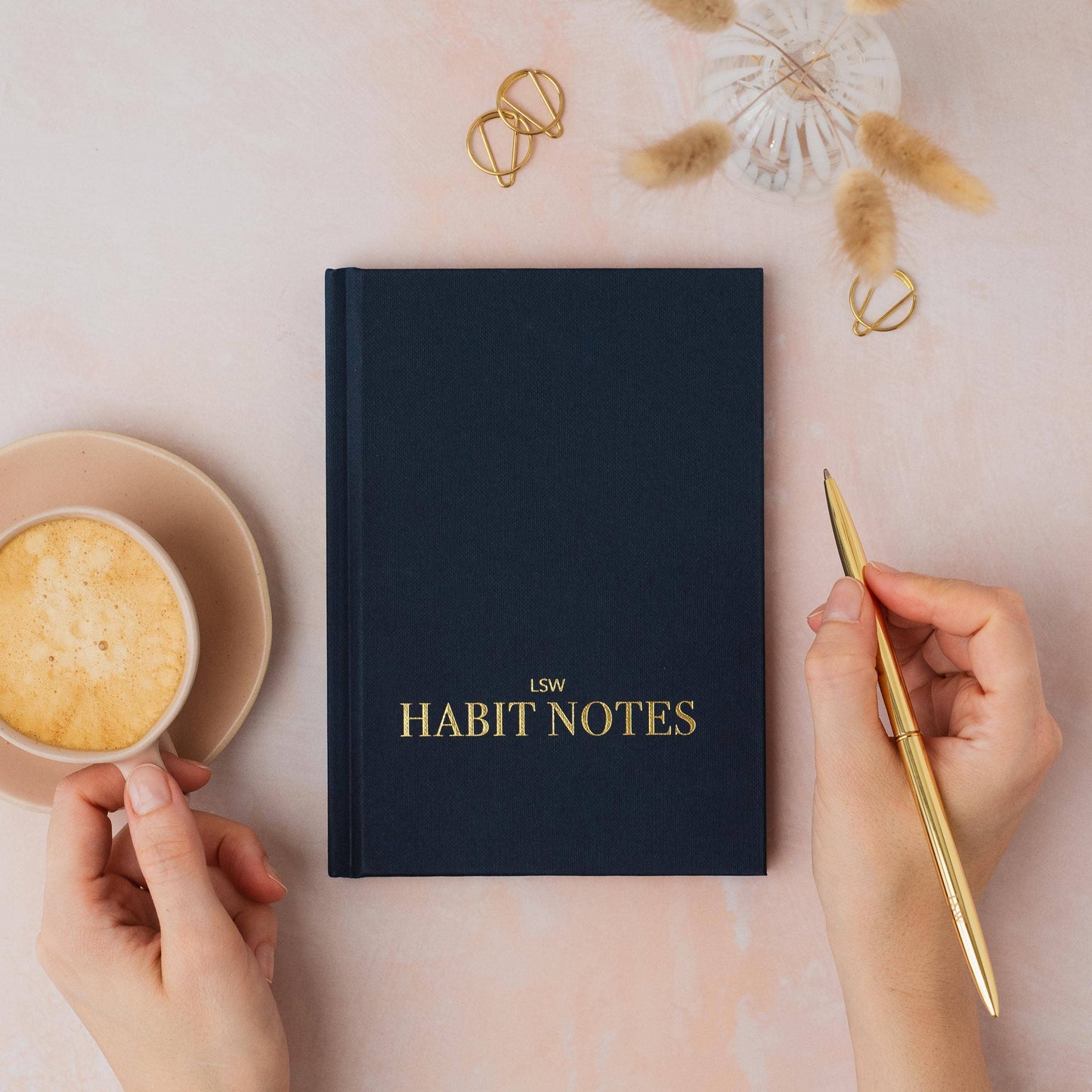 dark blue notebook titled 'LSW Habit Notes' on a pink surface with a hand holding a gold pen and a cup of coffee.