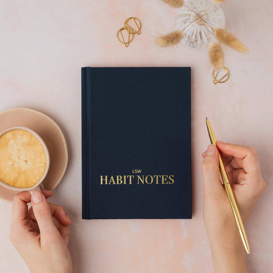 dark blue notebook titled 'LSW Habit Notes' on a pink surface with a hand holding a gold pen and a cup of coffee.