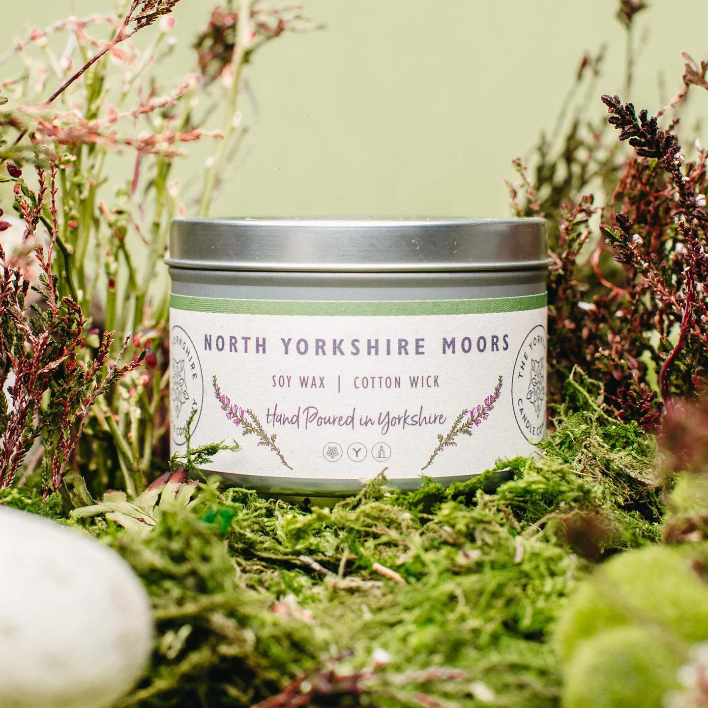 Candle in a tin labeled 'North Yorkshire Moors' surrounded by greenery
