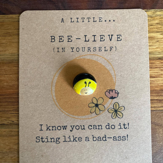 Button with bee design on a card with motivational text, placed on a wooden surface.