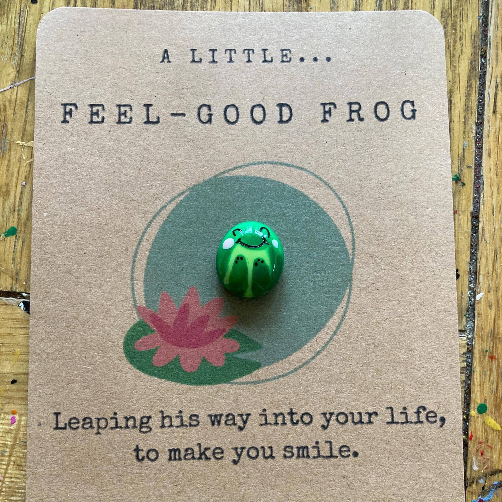 Green frog pin on a brown card with text and a pink flower illustration.