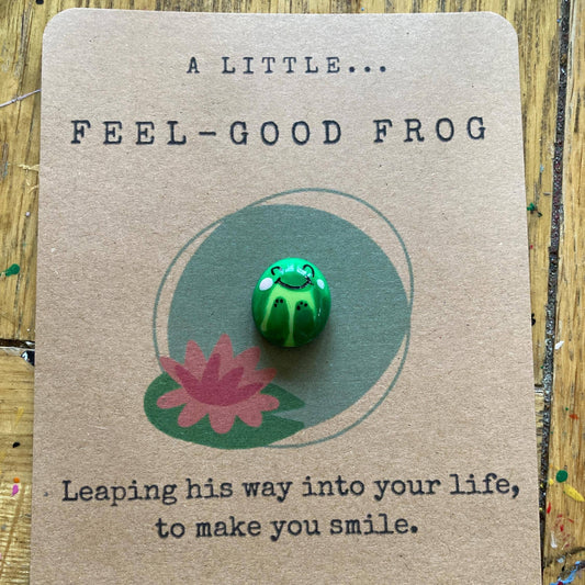 Green frog pin on a brown card with text and a pink flower illustration.