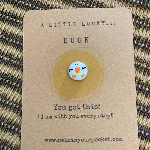 Pin with a duck design on a brown card with text, on a woven surface.