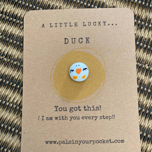 Pin with a duck design on a brown card with text, on a woven surface.