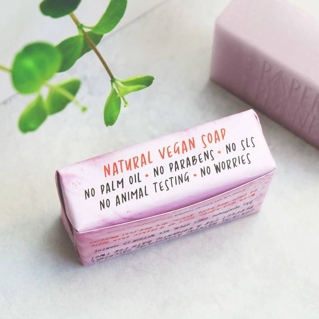 Pink soap bar with a label on a light surface with green leaves in the background