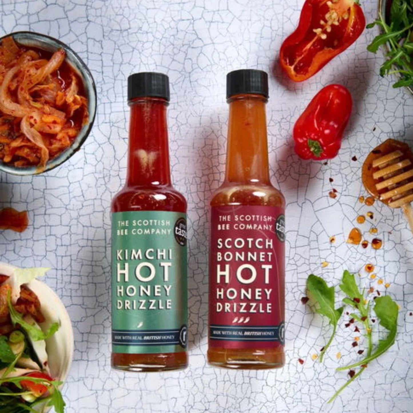 Two bottles of 'Kimchi Hot Honey Drizzle' and 'Scotch Bonnet Hot Honey Drizzle' from The Scottish Bee Company on a textured surface with food ingredients.