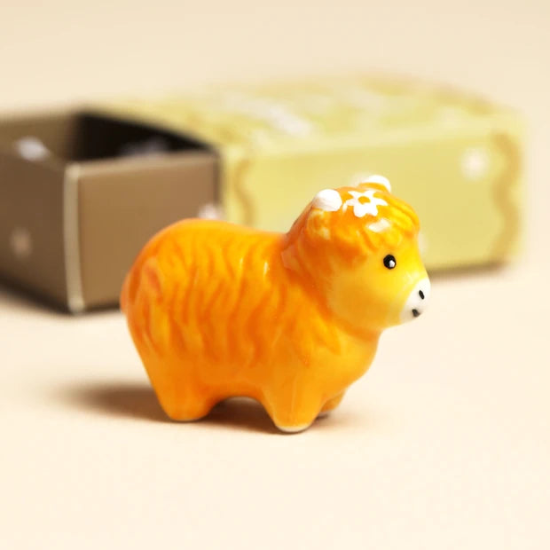 Tiny ceramic Highland cow token in warm amber glaze with a white flower on its head