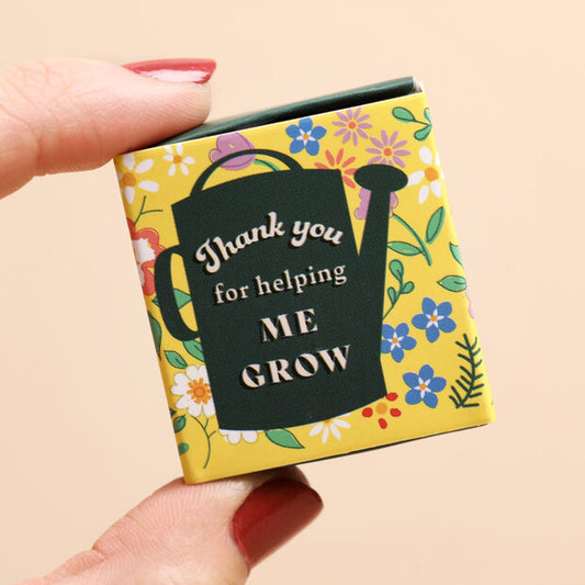 Small yellow box with floral design and text 'Thank you for helping ME GROW' held by a hand.