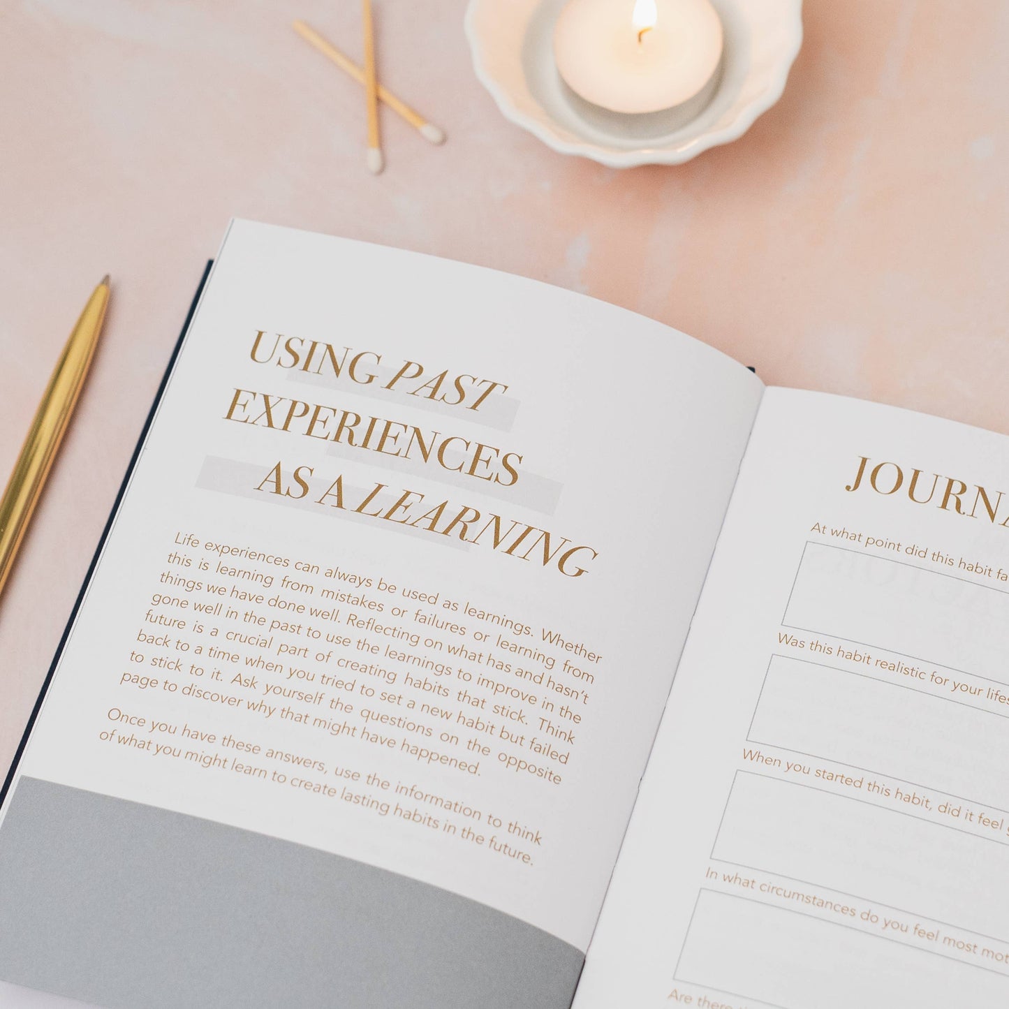 Open book with text on a soft pink background with candles and pens.